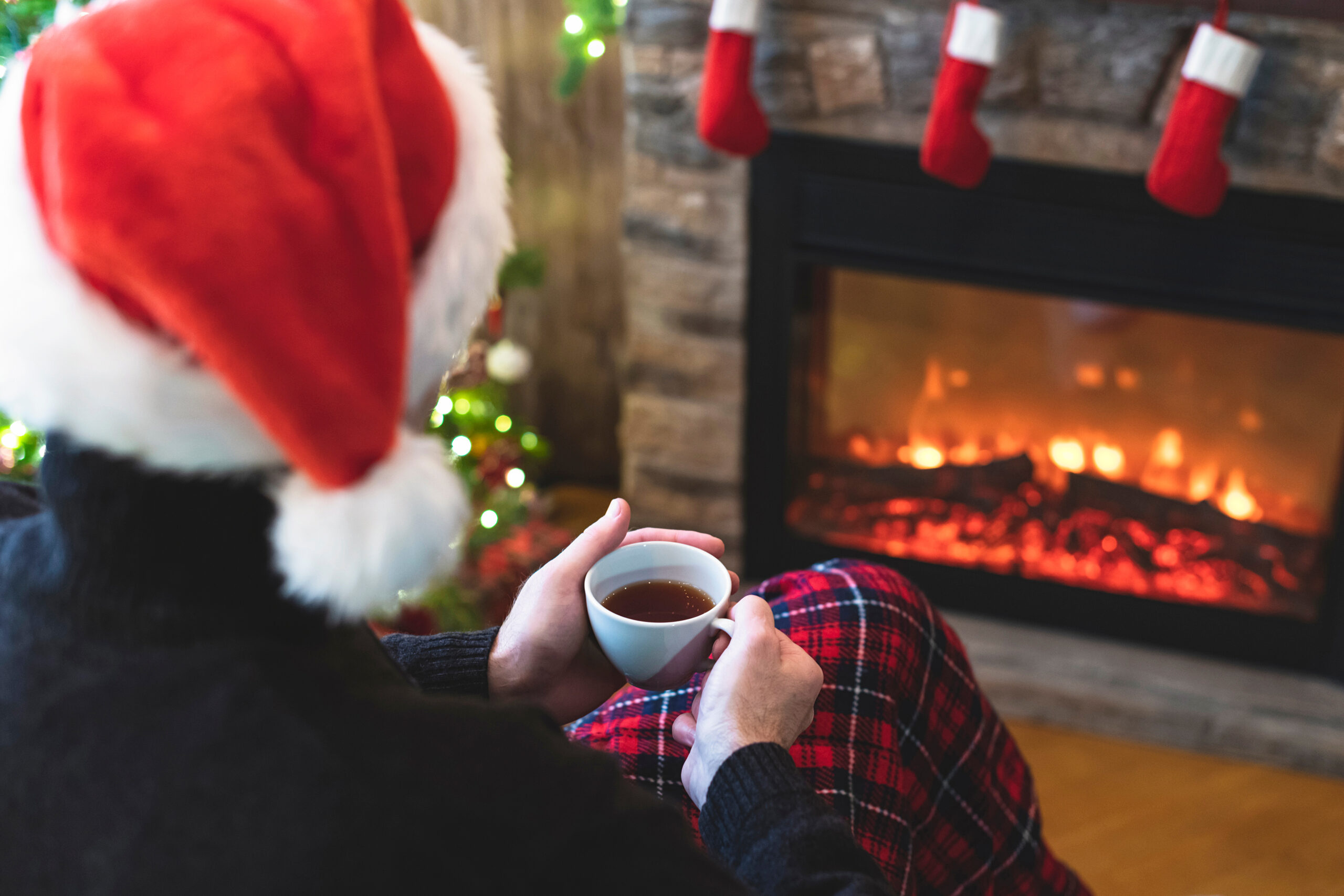 Person wearing a Santa hat holding a cup of tea, sitting by a cozy fireplace with holiday stockings and festive decorations, reflecting on managing holiday stress after divorce.
